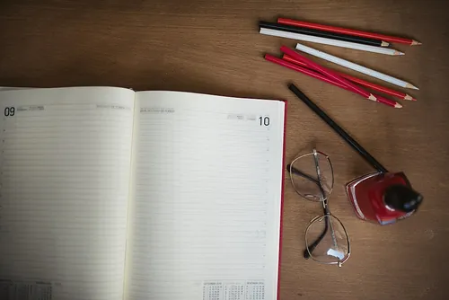 A planner is open to a new week with blank pages on a wooden desk. Nearby are colored pencils, glasses, and ink, suggesting an organized wor