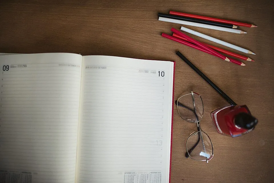 A planner is open to a new week with blank pages on a wooden desk. Nearby are colored pencils, glasses, and ink, suggesting an organized wor